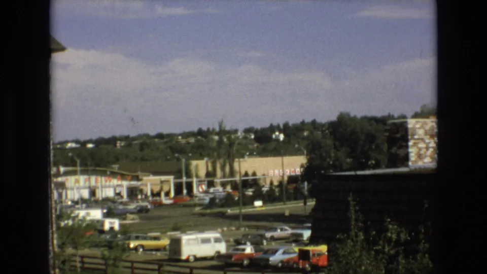 1973 Colorado Springs home-movie view through a dark window or balcony frame toward a roadside commercial area with parked classic cars and distant hills.