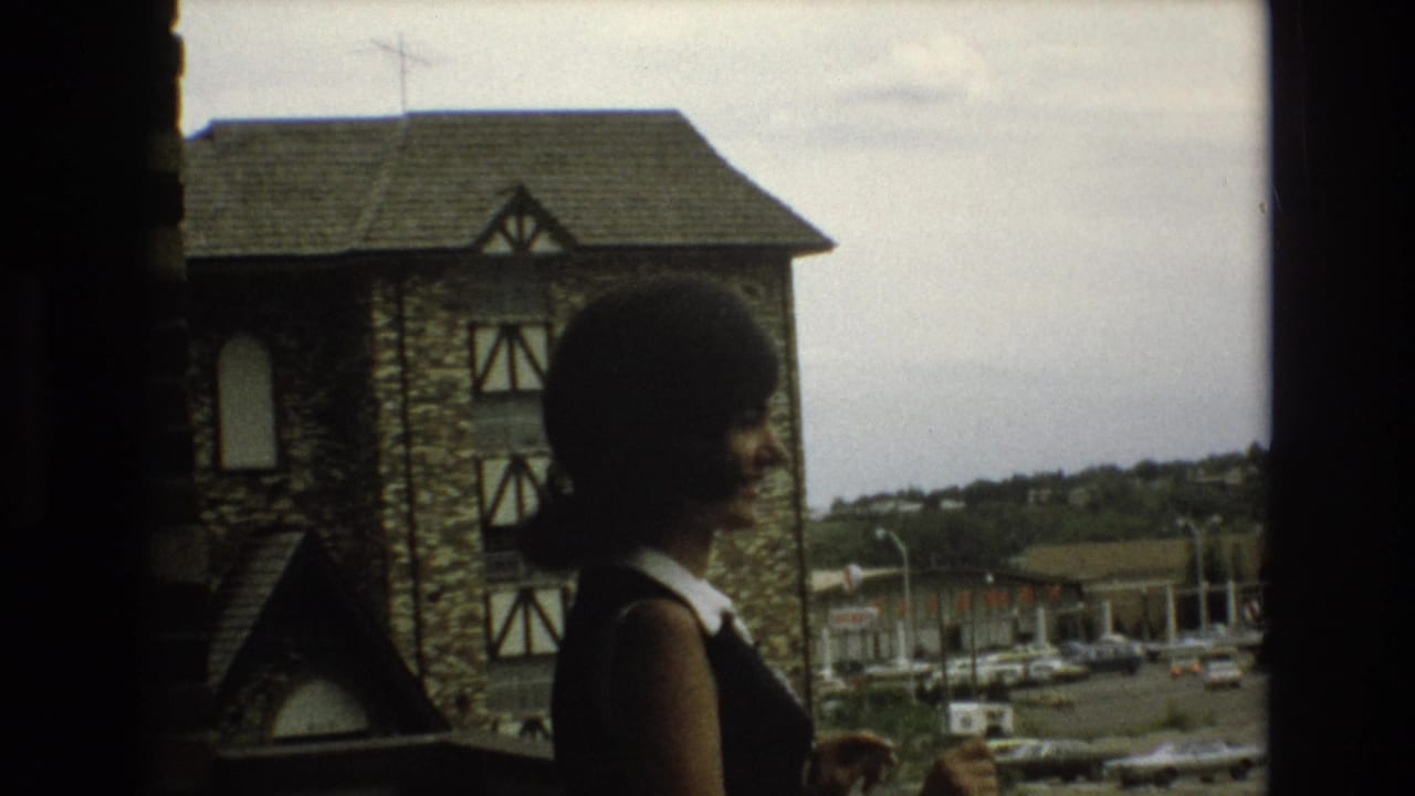 A woman with a dark bob haircut stands on an elevated walkway or balcony in Colorado Springs, with a stone half-timbered building and parked cars behind her.