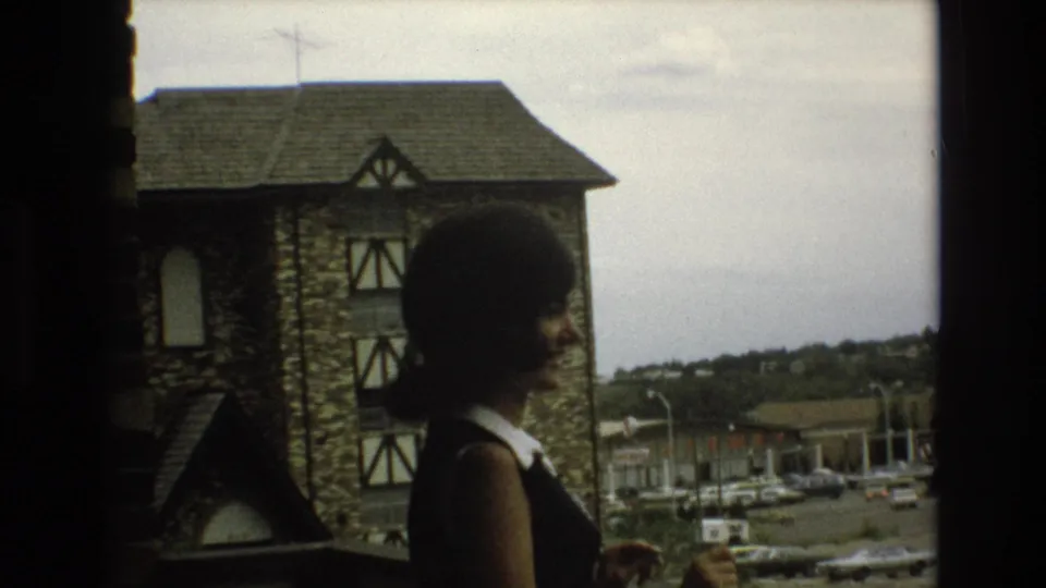 A woman with a dark bob haircut stands on an elevated walkway or balcony in Colorado Springs, with a stone half-timbered building and parked cars behind her.
