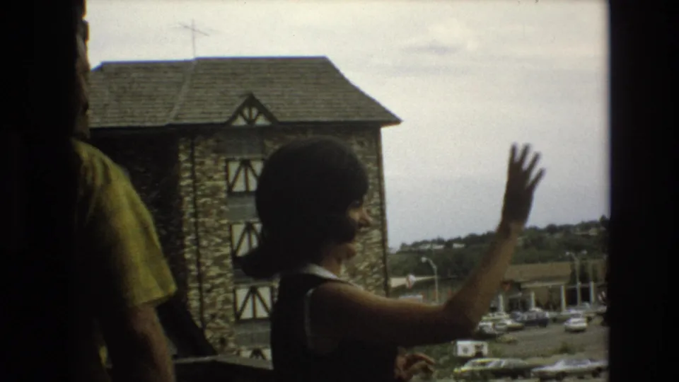 The woman raises her hand in a wave from the balcony or walkway, with a partially visible man in a yellow shirt and the stone building behind them.