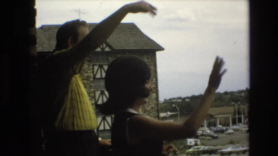 A man in a yellow shirt and a woman in a sleeveless dark dress wave toward the camera from an elevated balcony, with 1970s cars and roadside buildings in the background.