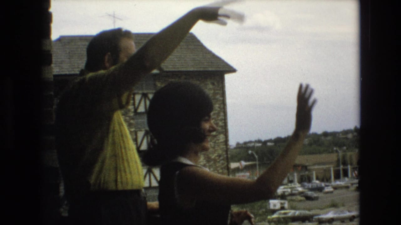 In 1973 Colorado Springs home-movie footage, an adult man in a yellow patterned shirt and an adult woman in a dark sleeveless dress gesture and wave from a window with a stone building and parked cars beyond.