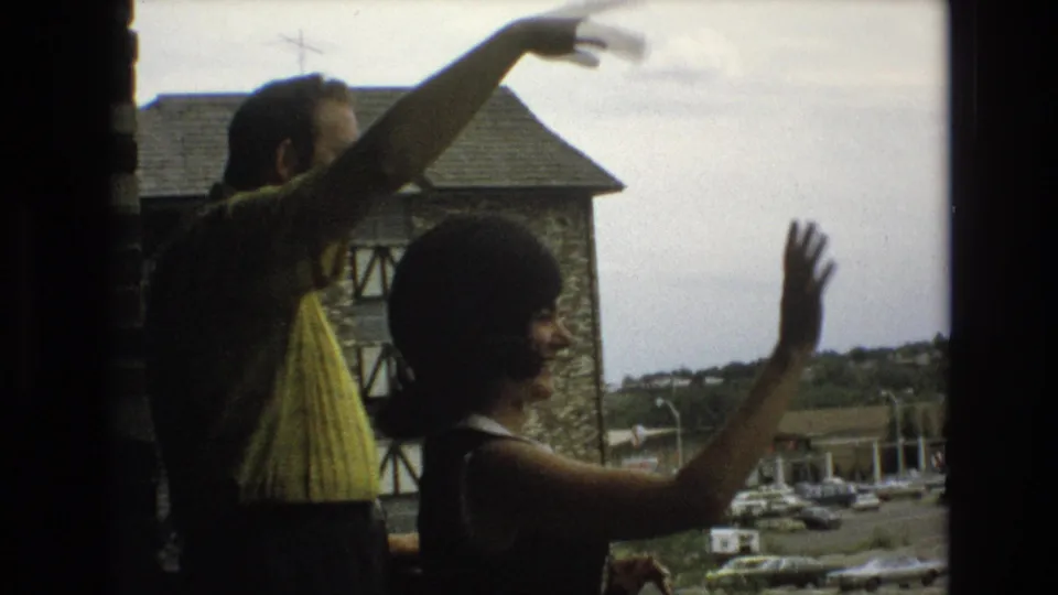 In 1973 Colorado Springs home-movie footage, an adult man in a yellow patterned shirt and an adult woman in a dark sleeveless dress gesture and wave from a window with a stone building and parked cars beyond.