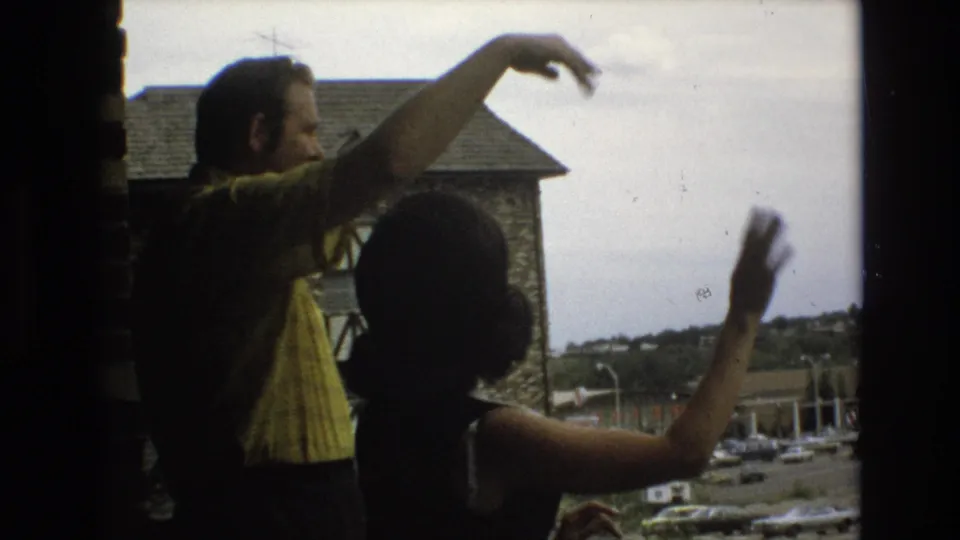 The man in a yellow patterned shirt extends his arm outward as the woman turns slightly away and waves, with the exterior building and parking area visible through the window.