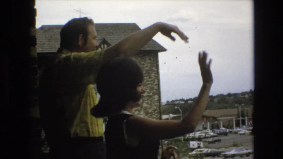 The two adults continue a goodbye gesture from the window, partly silhouetted against the outdoor view of a stone building, cars, and distant landscape.
