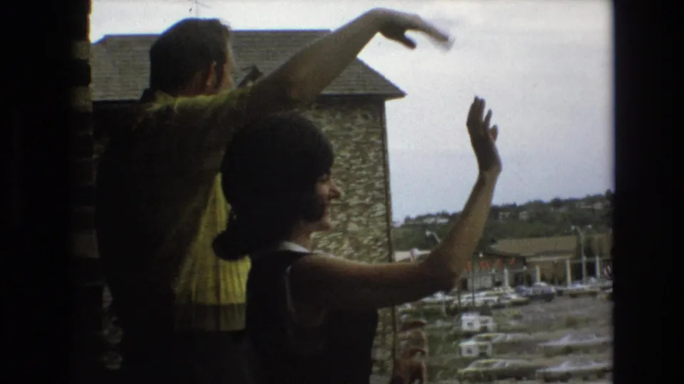 The woman smiles while waving from the window as the man stands behind her with his arm raised, framed by soft 8mm exposure and an outdoor parking area beyond.