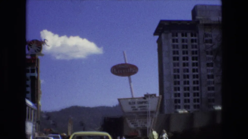 1973 Lake Tahoe street view with the Tropicana hotel sign, a marquee, passing cars, pedestrians, a tall building, mountains, and blue sky.
