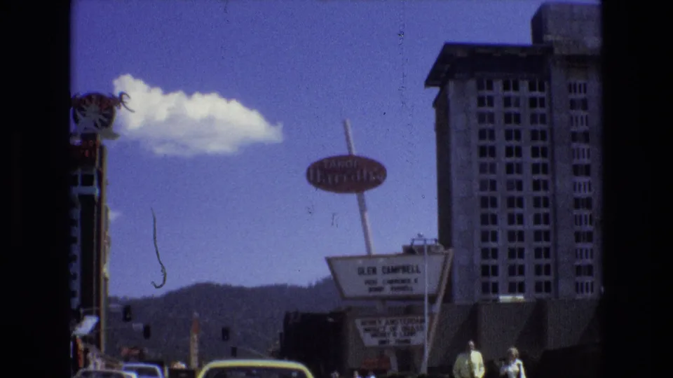 Lake Tahoe, Nevada street frame showing the Tropicana sign, hotel marquee, pedestrians near the sidewalk, cars in traffic, distant mountains, and a tall building.