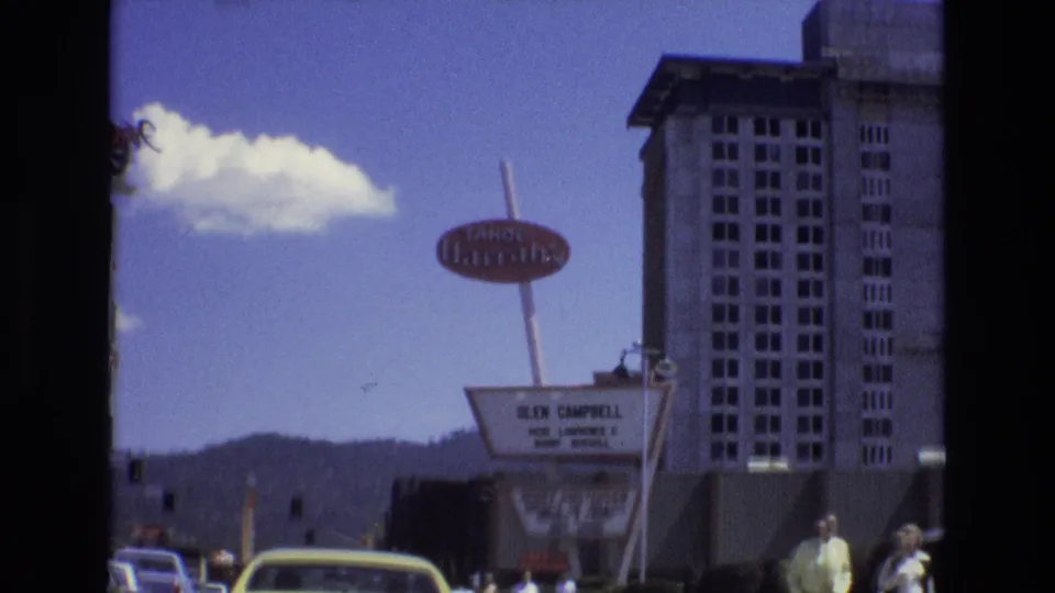 1973 Lake Tahoe street view with a yellow car in the foreground, the Tropicana sign above, a marquee listing performers, pedestrians, and resort-area buildings.