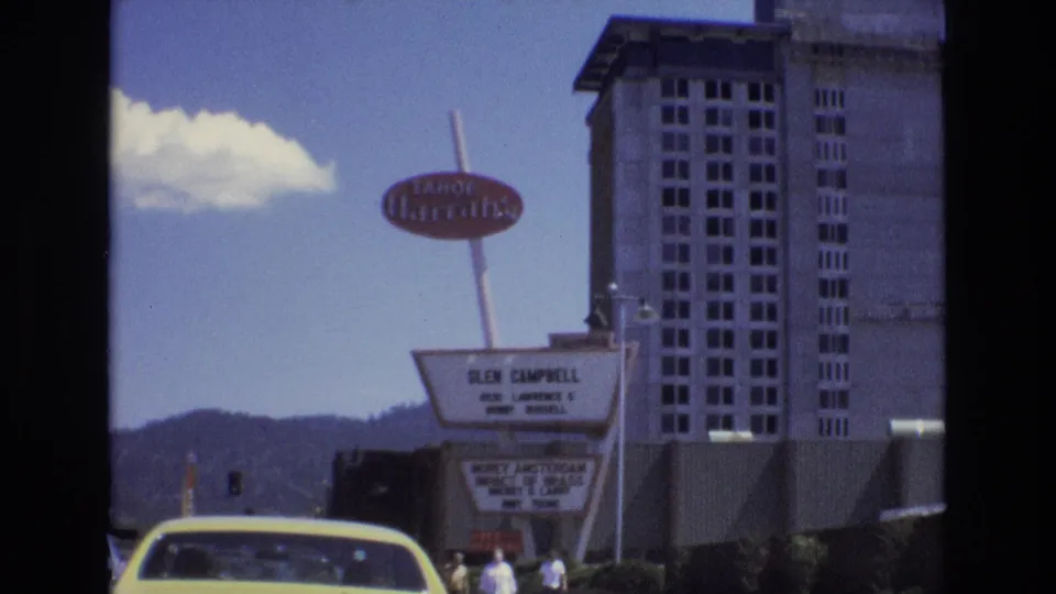 Lake Tahoe 1973 frame showing the Tropicana roadside sign, hotel marquee, yellow car, several pedestrians, traffic lights, a tall building, mountains, and a bright cloud.