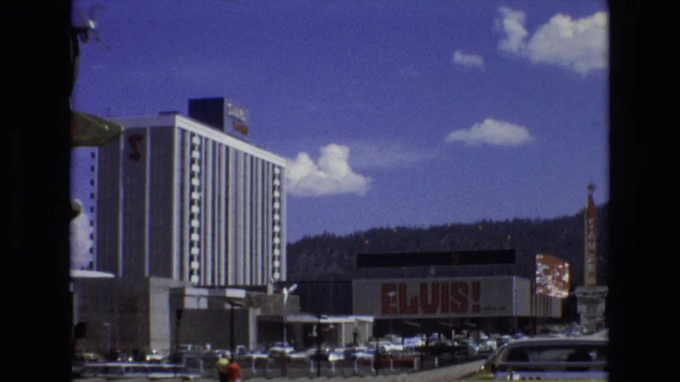 1973 Lake Tahoe, Nevada frame showing a tall resort tower, large ELVIS signage, parked cars, roadside signs, and wooded hills under bright sky.