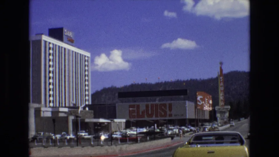 1973 Lake Tahoe resort-area home-movie frame with a large ELVIS sign, hotel tower, car-filled street edge, and mountain landscape.