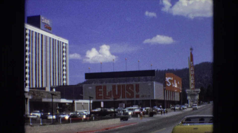 1973 Lake Tahoe, Nevada view of a roadside building with a prominent Elvis advertisement, a tall hotel tower on the left, cars along the curb, and pedestrians crossing or walking nearby.