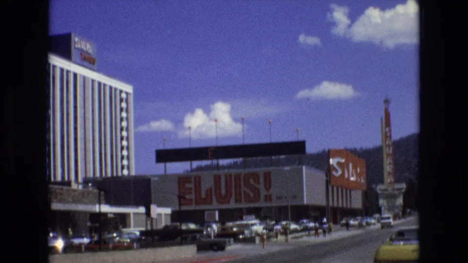 1973 Lake Tahoe, Nevada street frame showing the Elvis advertisement, nearby Sahara sign, hotel facade, moving and parked cars, distant pedestrians, and forested hills under a blue sky.
