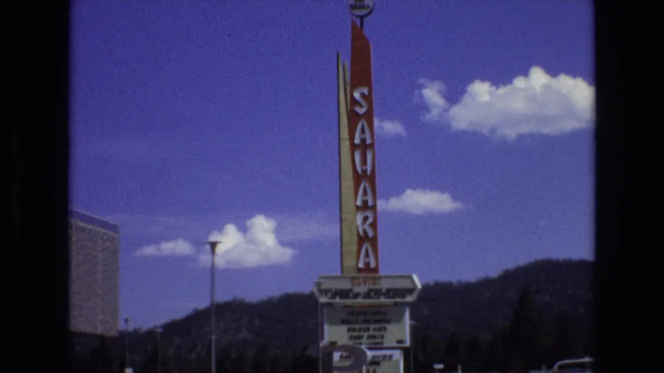1973 Lake Tahoe, Nevada home-movie frame centered on the Sahara vertical sign and marquee, with a streetlight, mountain ridge, and blue sky behind it.