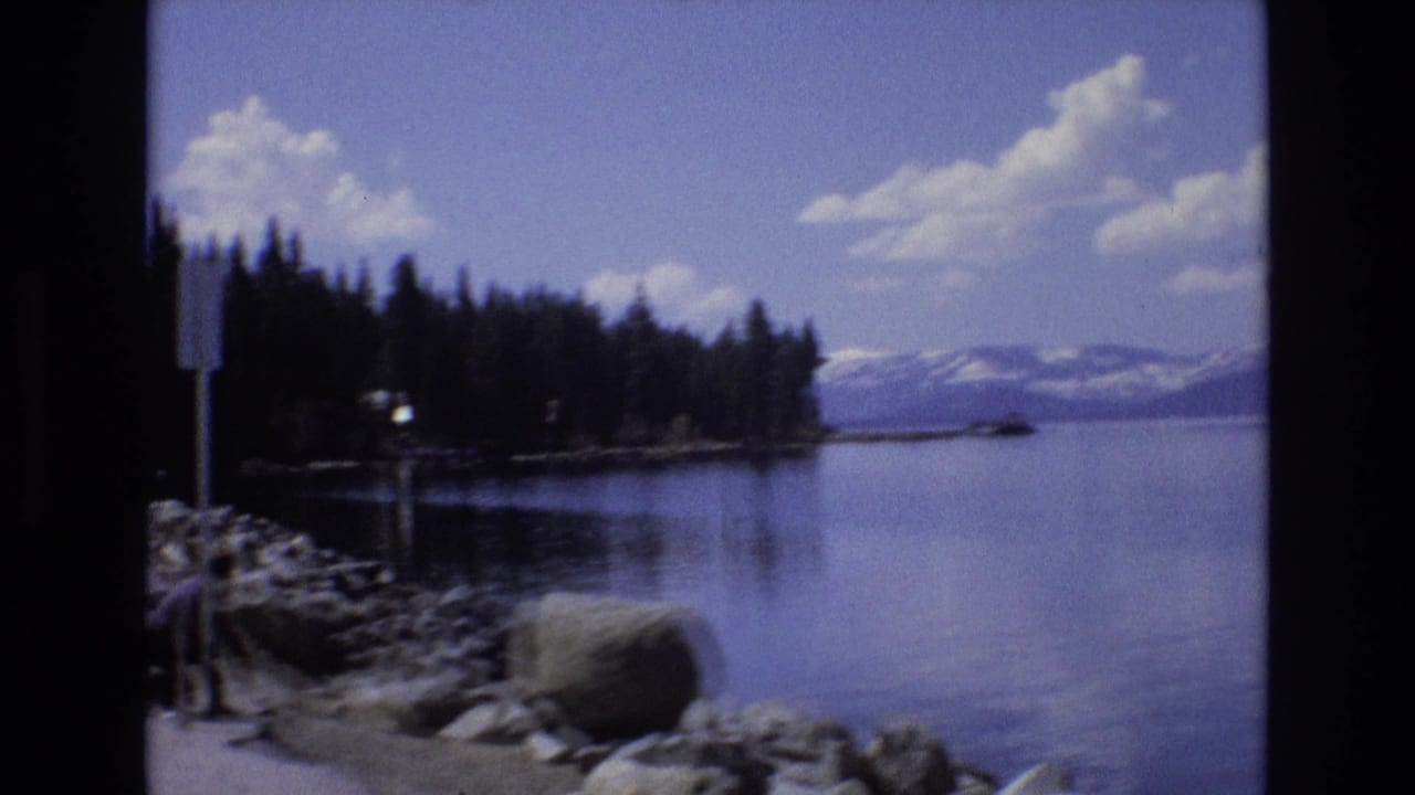 1973 Lake Tahoe, Nevada home-movie frame looking across a rocky shoreline, evergreen trees, calm water, and snow-capped mountains in the distance.