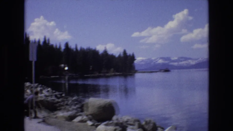 1973 Lake Tahoe, Nevada home-movie frame looking across a rocky shoreline, evergreen trees, calm water, and snow-capped mountains in the distance.