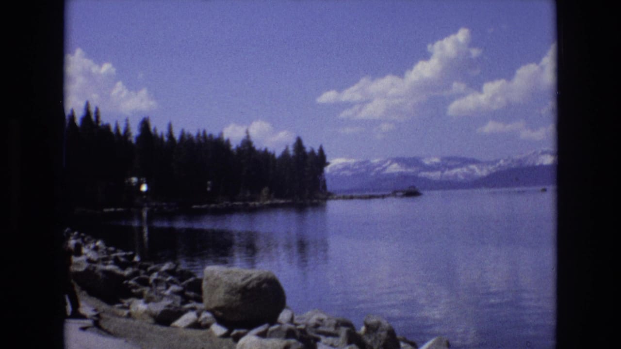 1973 Lake Tahoe, Nevada home-movie frame with shoreline rocks in the foreground, a forested point, calm lake water, and snow-capped Sierra mountains beyond.