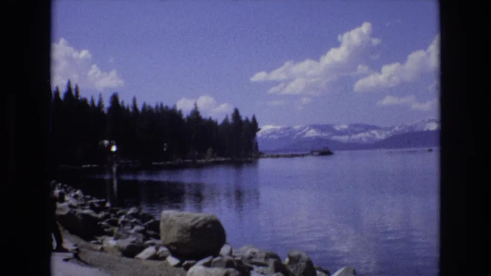 1973 Lake Tahoe, Nevada home-movie frame with shoreline rocks in the foreground, a forested point, calm lake water, and snow-capped Sierra mountains beyond.