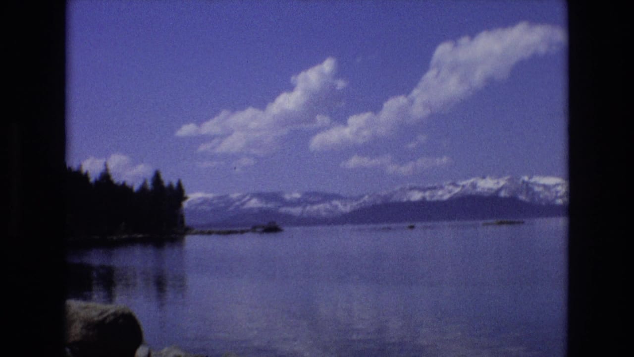 1973 Lake Tahoe, Nevada home-movie frame showing a wide lake view with a dark evergreen shoreline at left, open water, bright clouds, and snow-capped mountains.