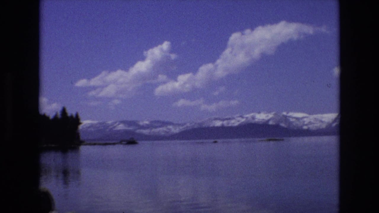 Wide lake with snow-capped mountains, blue sky, clouds, and a dark shoreline at left.