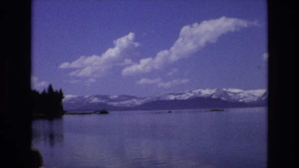 1973 Lake Tahoe, Nevada home-movie frame showing a reflective lake beneath snow-capped mountains and scattered clouds, with dark trees or shoreline along the left edge.