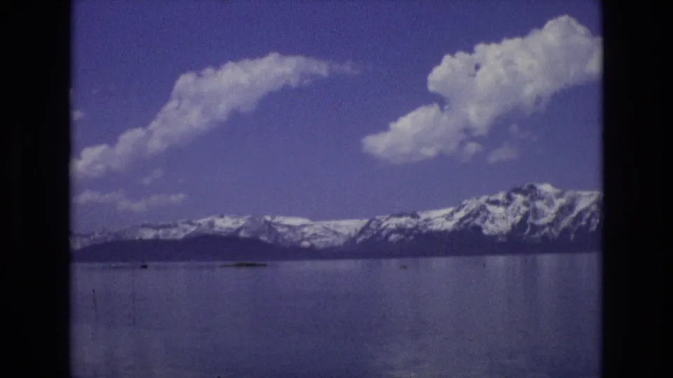 1973 Lake Tahoe, Nevada home-movie view of a reflective lake and snowy mountain ridge, with large white clouds overhead and small distant shapes near the waterline.