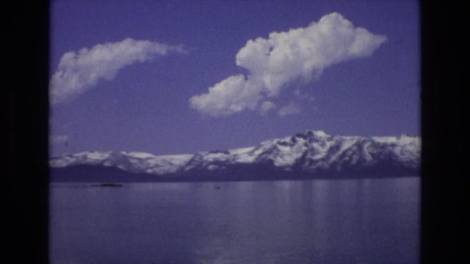 1973 Lake Tahoe, Nevada frame showing a broad lake foreground, snow-covered mountains on the horizon, and a prominent white cloud formation against blue sky.