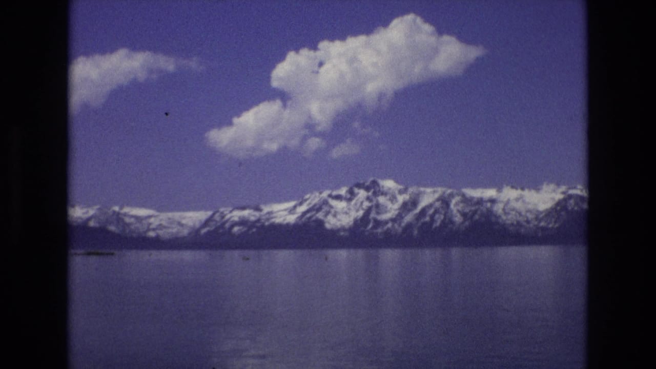 1973 Lake Tahoe, Nevada home-movie frame showing calm water, distant snow-capped mountains, and a large white cloud under a blue sky.