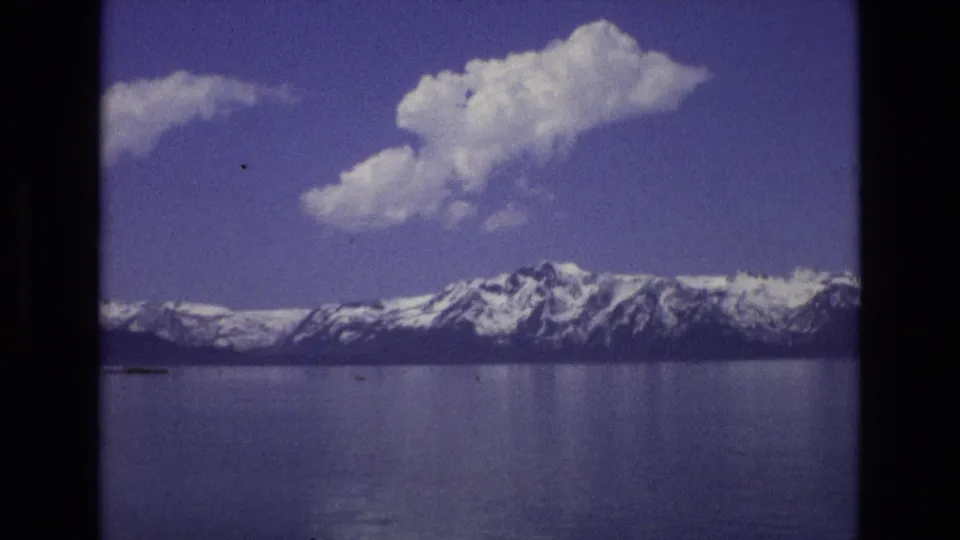 1973 Lake Tahoe, Nevada home-movie frame showing calm water, distant snow-capped mountains, and a large white cloud under a blue sky.