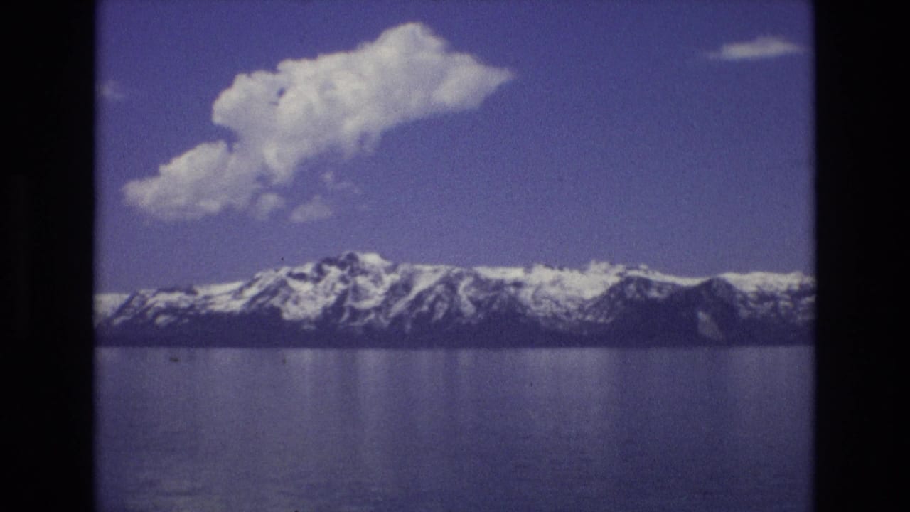 Snowy mountain range across still water under blue sky with clouds.