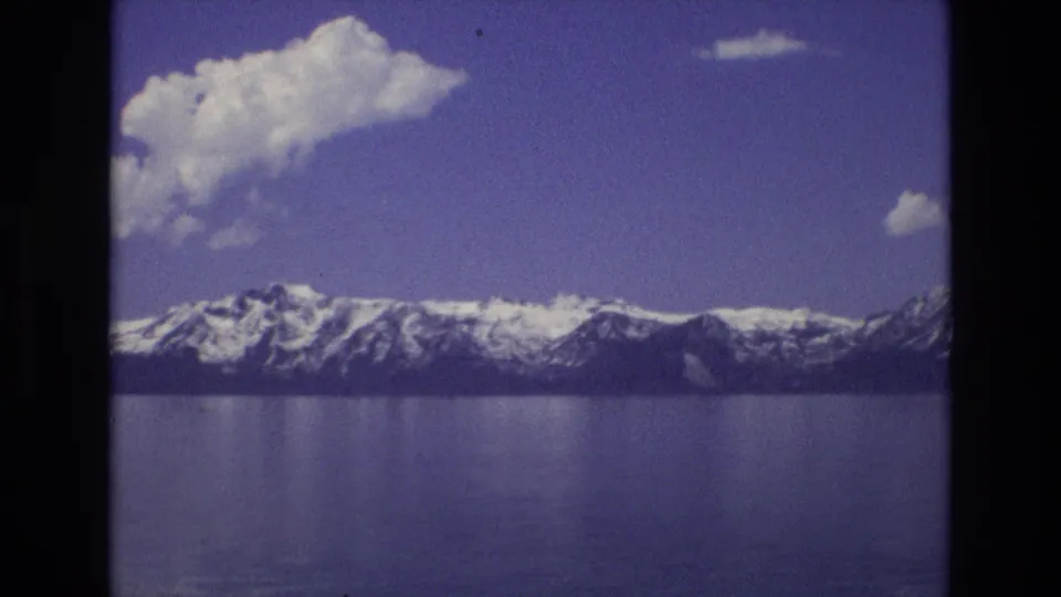 1973 Lake Tahoe, Nevada home-movie view of a snowy ridgeline across calm water, with a large cloud drifting over the mountains.