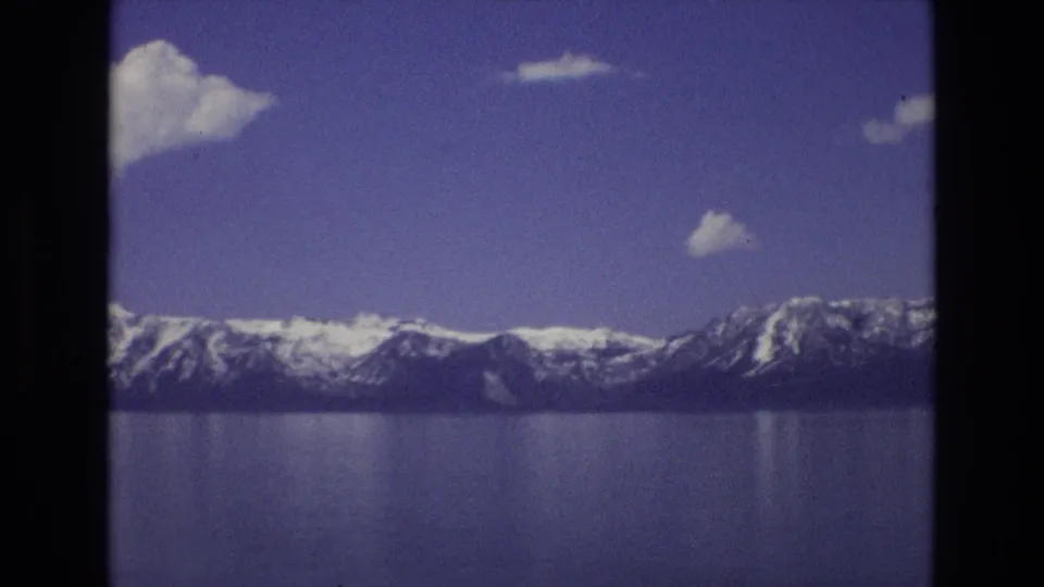 1973 Lake Tahoe, Nevada frame showing distant snow-capped mountains and calm reflective water beneath a mostly clear blue sky with small clouds.