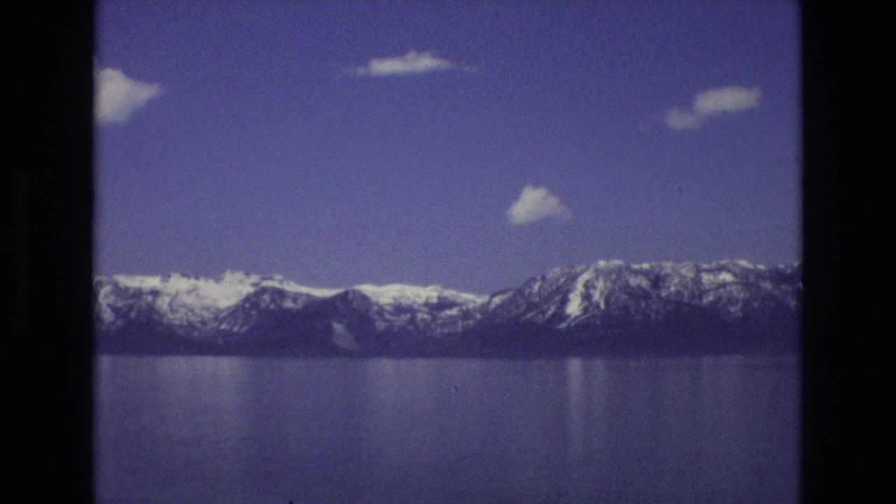 Snow-capped mountains across a calm blue lake under a clear sky with small clouds.