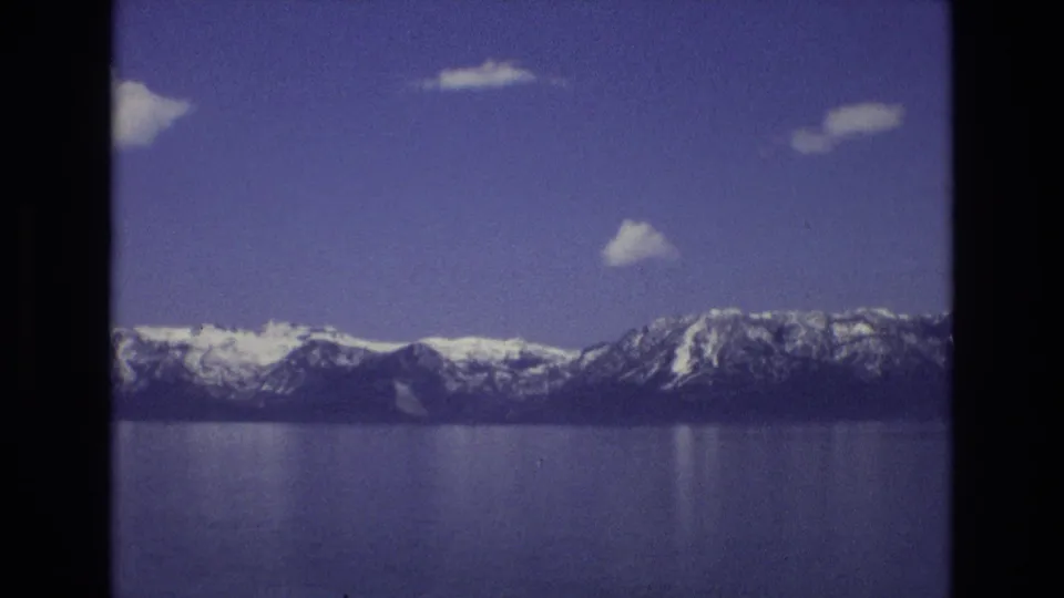 1973 8mm home-movie view of Lake Tahoe, Nevada, with snow-capped mountains across calm blue water and small white clouds in a purple-blue sky.