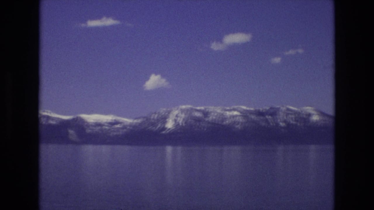 1973 Lake Tahoe home-movie frame showing a distant snow-lined mountain range, still blue water, soft reflections, and scattered clouds within the rounded 8mm frame.