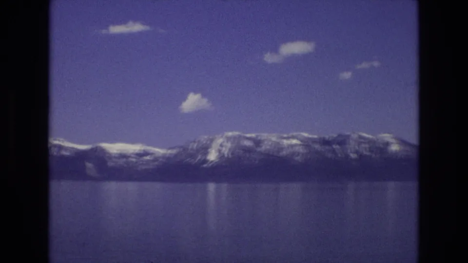 1973 Lake Tahoe home-movie frame showing a distant snow-lined mountain range, still blue water, soft reflections, and scattered clouds within the rounded 8mm frame.