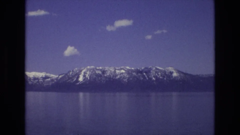 1973 8mm view at Lake Tahoe with a snow-streaked mountain ridge, broad lake surface, scattered clouds, and visible home-movie grain and softness.