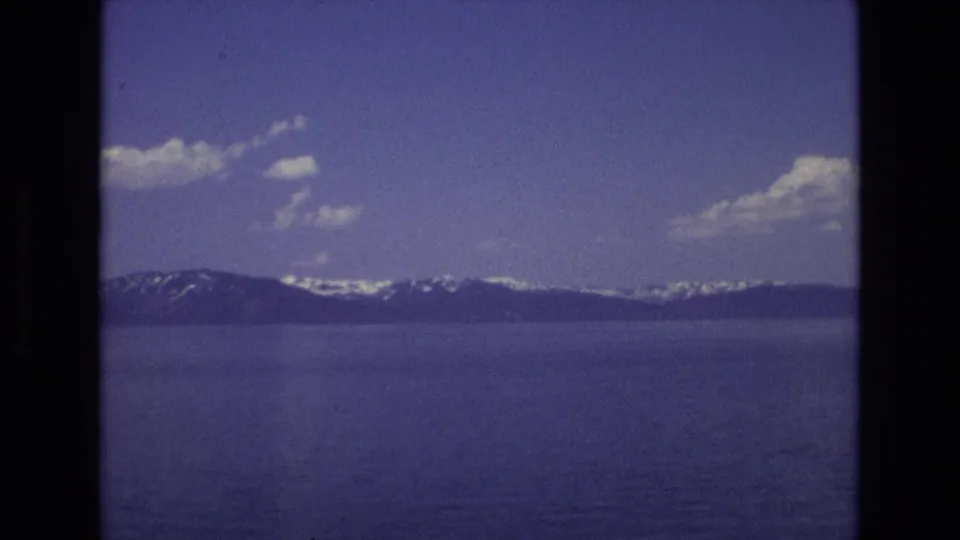 1973 Lake Tahoe frame showing a wider blue lake view, distant mountain ridges with patches of snow, scattered clouds, and a small dark sky speck that may be a bird, aircraft, or film artifact.