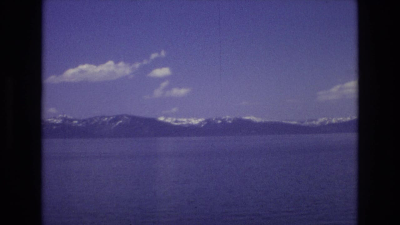1973 Lake Tahoe home-movie frame showing blue lake water, a dark mountain ridge with snow patches, and scattered white clouds in a purple-tinted 8mm scan.