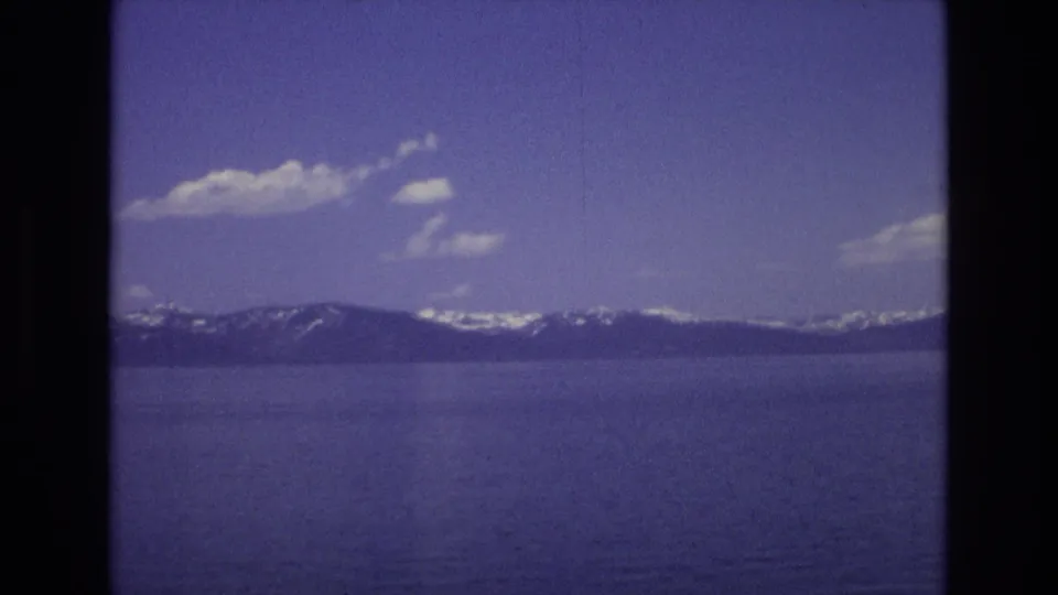 1973 Lake Tahoe home-movie frame showing blue lake water, a dark mountain ridge with snow patches, and scattered white clouds in a purple-tinted 8mm scan.