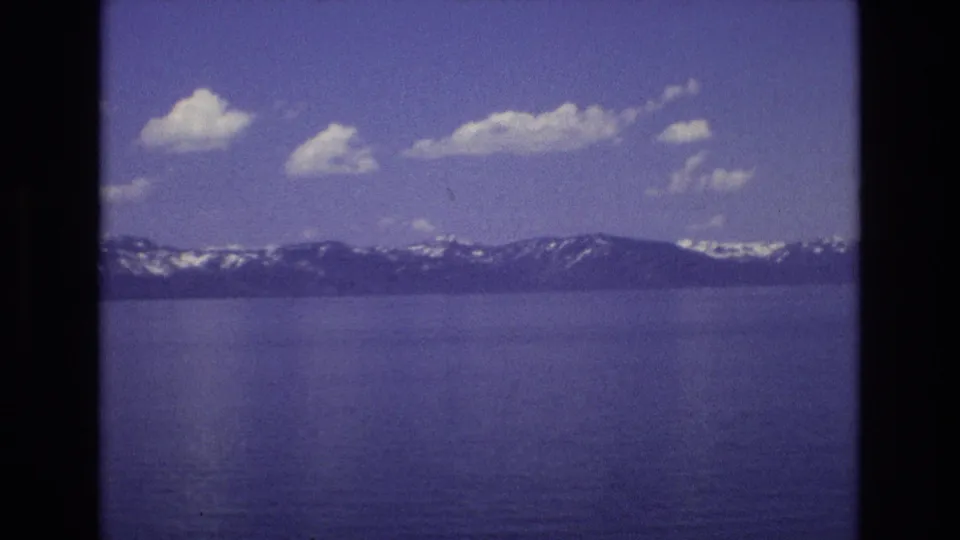 1973 home-movie frame of Lake Tahoe with rippled blue water, a snow-streaked mountain ridge, scattered clouds, and a small dark speck over the scene that may be film debris or a distant object.