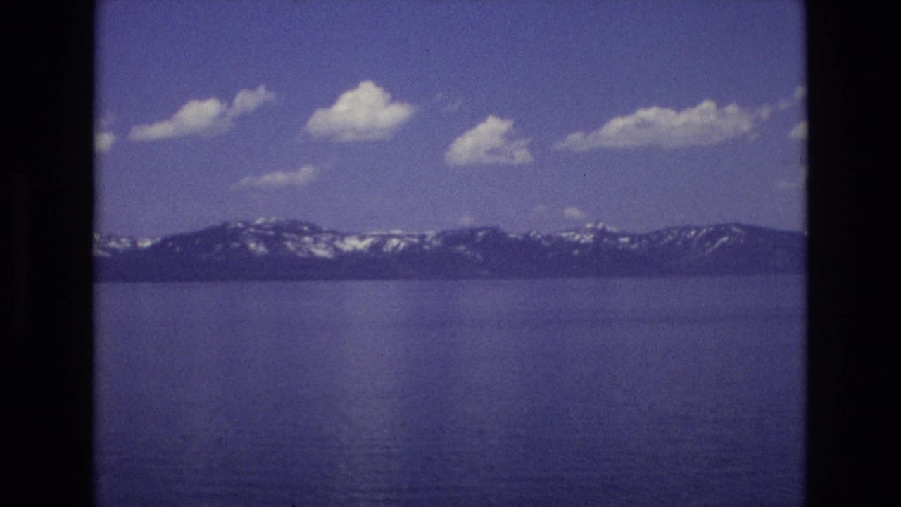 1973 Lake Tahoe home-movie frame showing deep blue water, distant snow-streaked mountains, scattered white clouds, and dark 8mm scan borders.