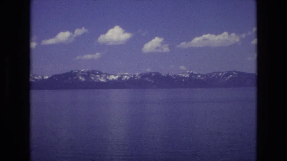 1973 Lake Tahoe home-movie frame showing deep blue water, distant snow-streaked mountains, scattered white clouds, and dark 8mm scan borders.