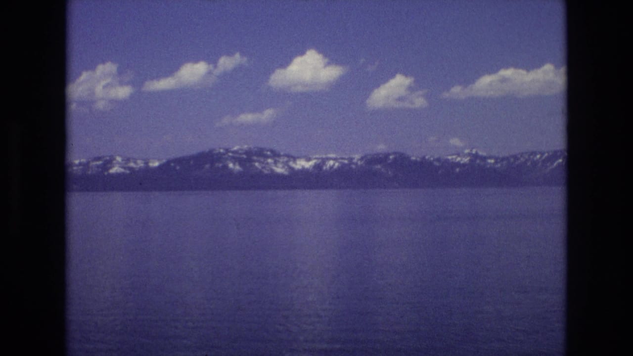 1973 Lake Tahoe frame looking across calm blue water toward a dark mountain ridge with snow patches and small white clouds overhead.