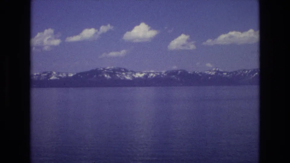 1973 Lake Tahoe frame looking across calm blue water toward a dark mountain ridge with snow patches and small white clouds overhead.