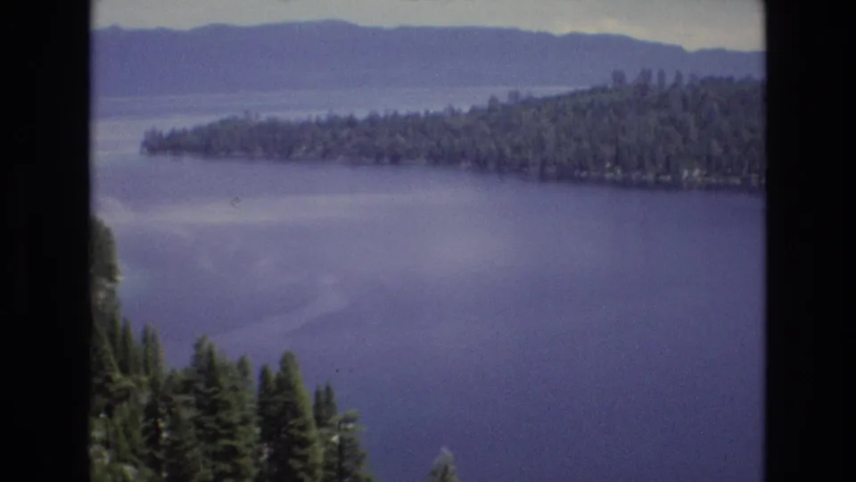 1973 Lake Tahoe home-movie view from above the water, with a forested shoreline, hazy distant hills, and dark evergreen trees at the lower edge.