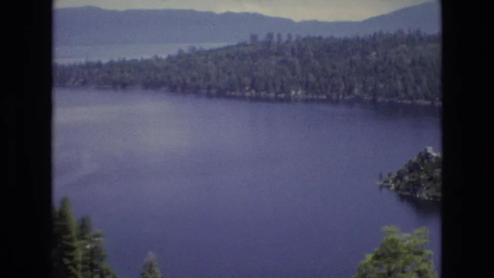 1973 Lake Tahoe frame showing blue water, a tree-covered shoreline, distant hills, and a small rocky island or point near the lower right corner.