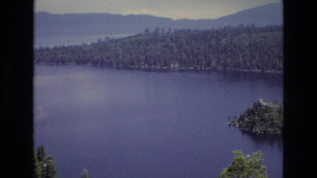 1973 8mm view of Lake Tahoe, Nevada with deep blue water, a small tree-covered island, forested shoreline, hazy mountains, and dark frame borders.