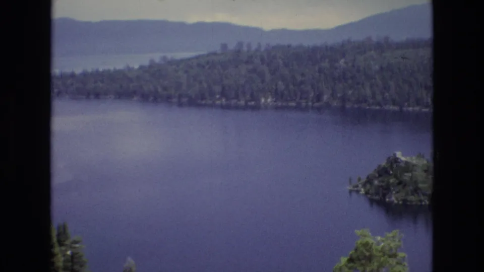 1973 8mm view of Lake Tahoe, Nevada with deep blue water, a small tree-covered island, forested shoreline, hazy mountains, and dark frame borders.